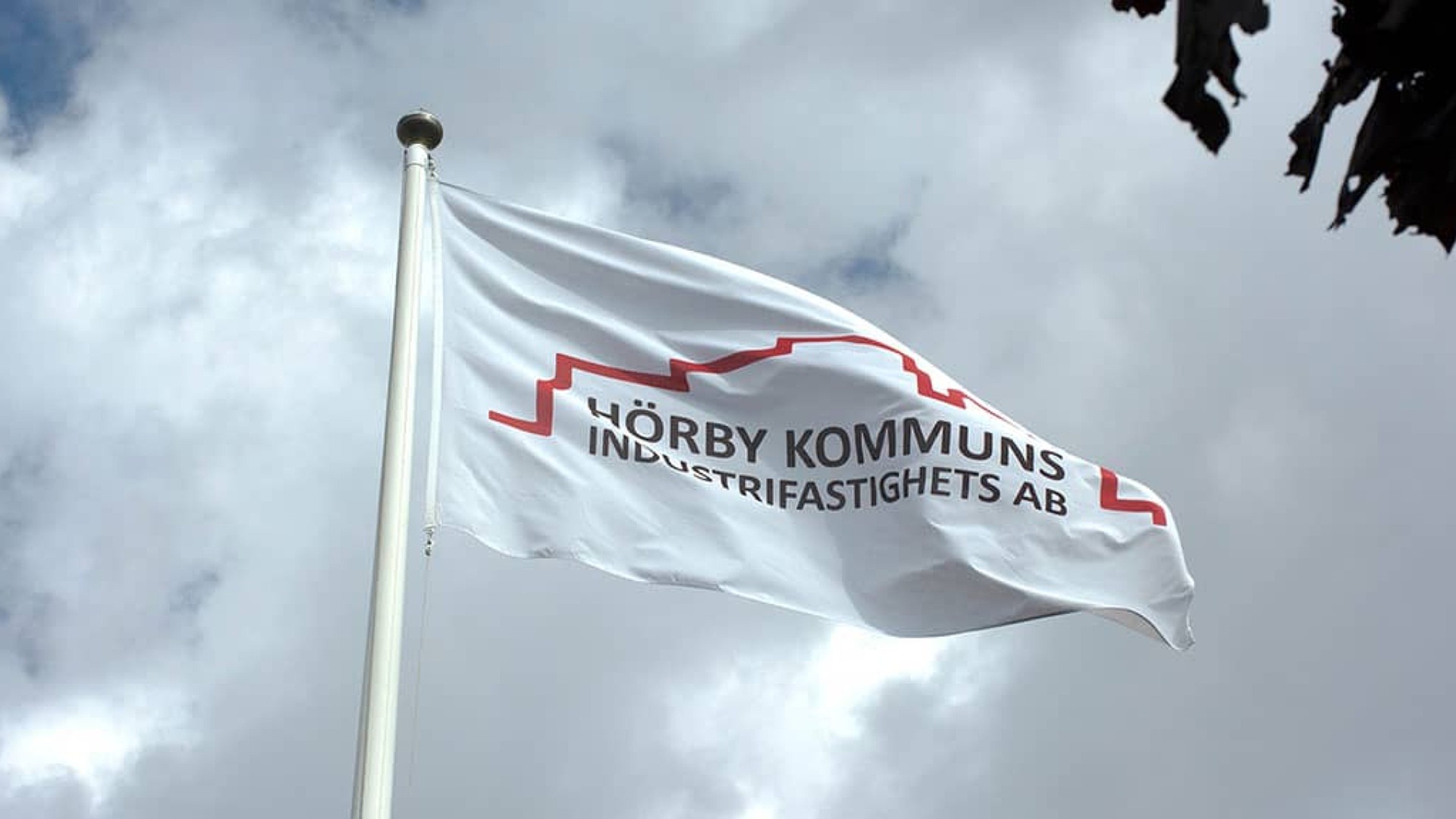 Vit flagga med Hörby Industrifastighets AB:s logotyp vajar på flaggstång