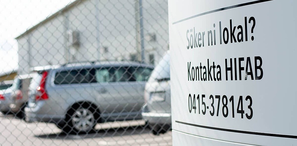 Skylt med texten "Söker ni lokal" i förgrunden. I bakgrunden finns en industrifastighet och parkerade bilar.